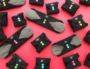 Reusable Sanitary Pads by Floating Lotus.  Award-winning Period Pads for eco periods, shown in mixed sizes small, medium, large and XL for every period. Some pads are open, others folded closed, set to a dark pink background.